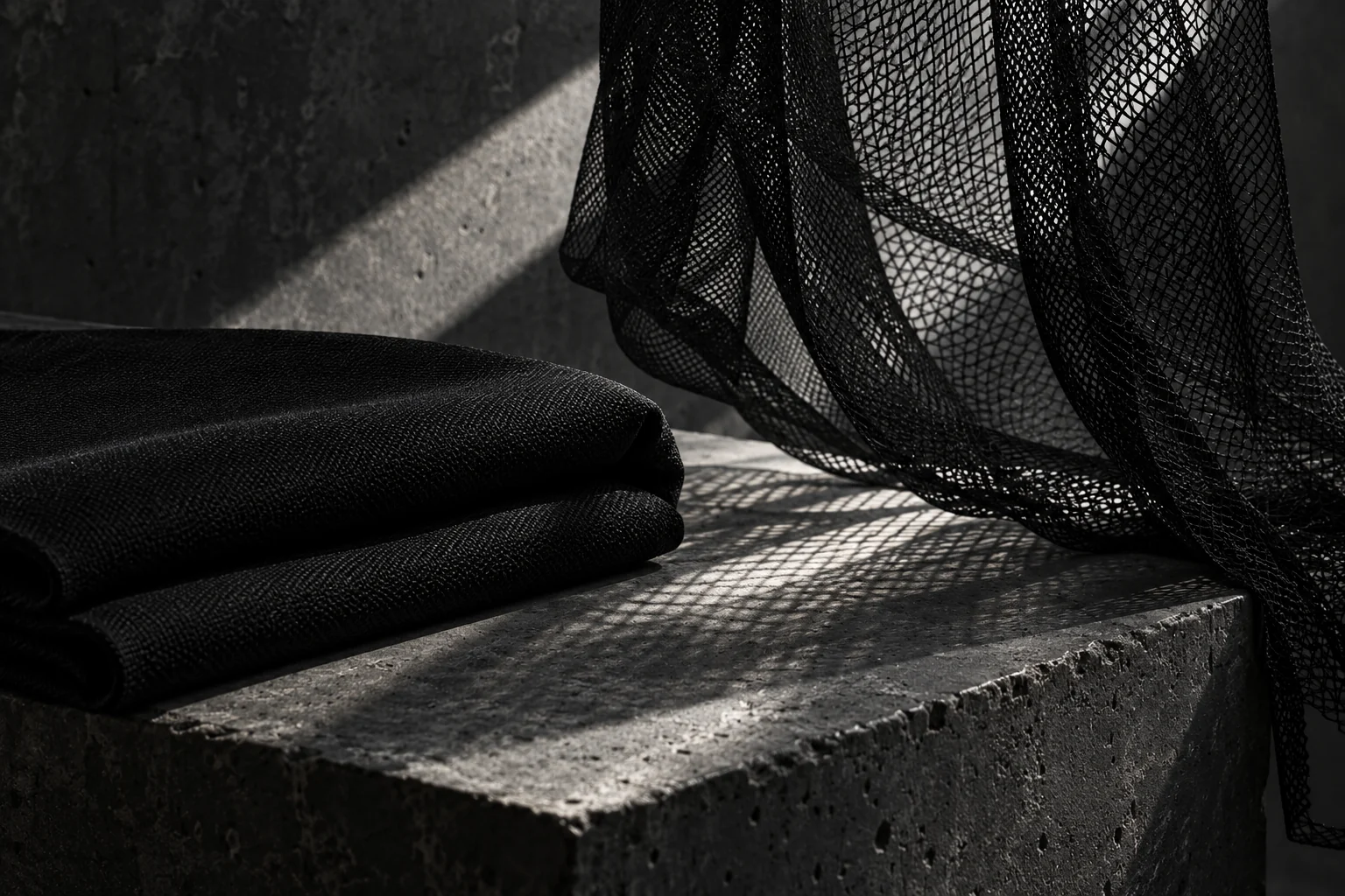 Plain black mesh casting a fine shadow beside a folded matte black textile on concrete