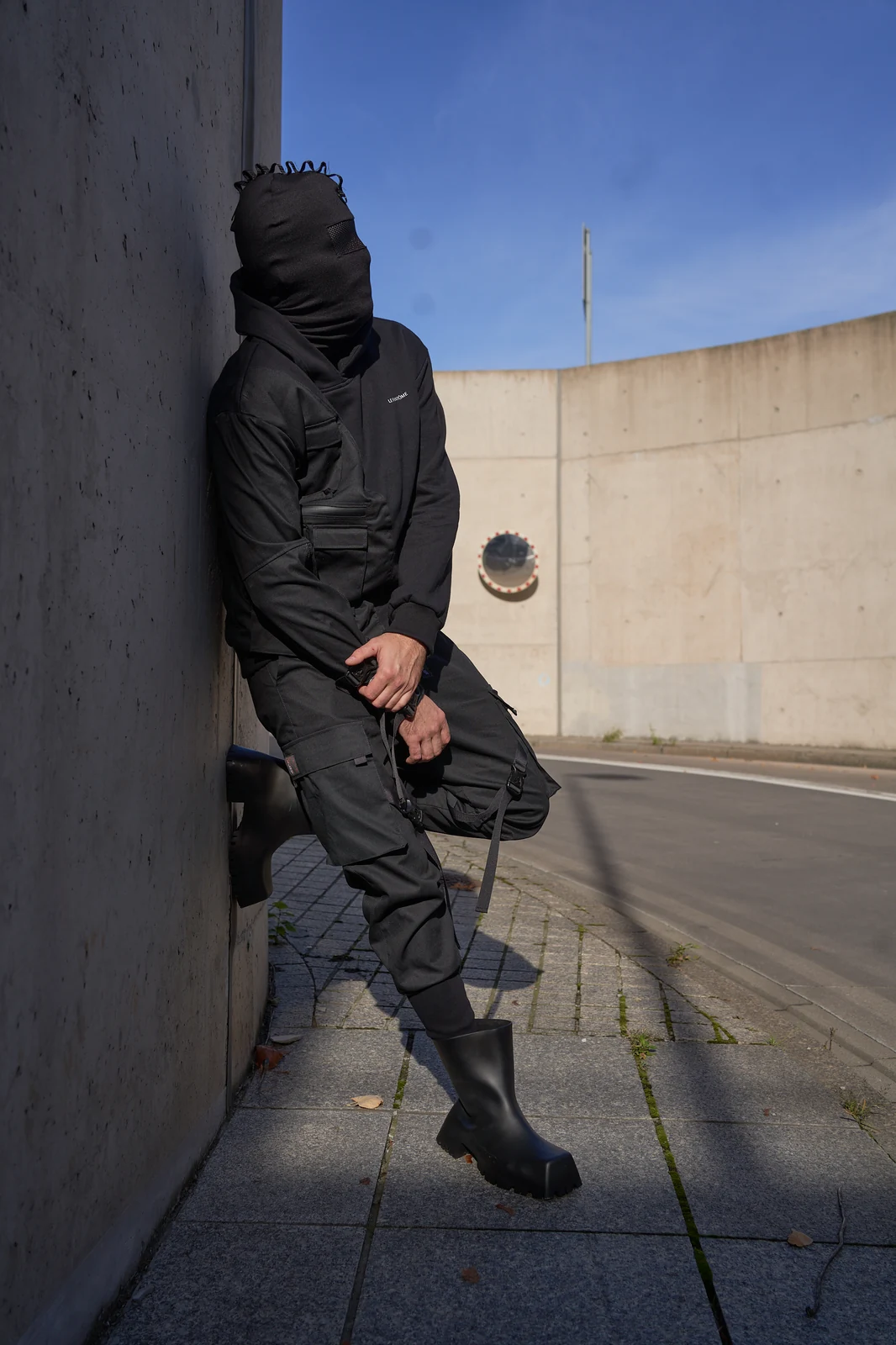 A layered black LF campaign outfit with a hood, face covering, cargo pockets and hanging straps, photographed beside a concrete wall